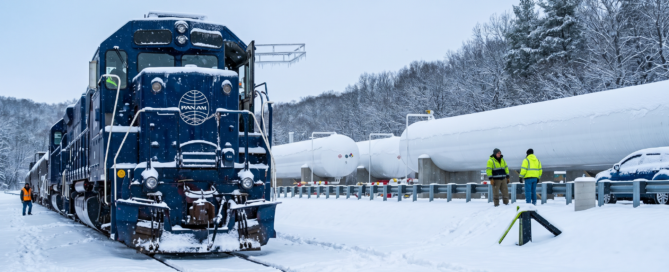 Blue Pan Am locomotive in a snow-covered rail yard with two workers in bright jackets nearby.