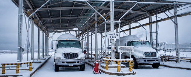 Snow-covered loading bays with two white trucks under a metal canopy; icicles hang from the roof in a cold industrial site.